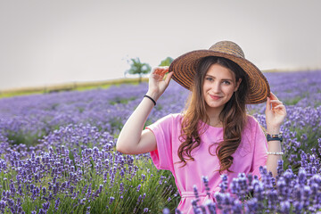 Beautiful brunette smiling girl in straw hat and pink dress is posing in purple lavender field.  Horizontally. 