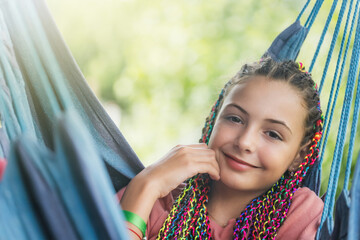 Closeup portrait of smiling young girl with colorful braids in her hair posing on blue hammock.  Horizontally. 