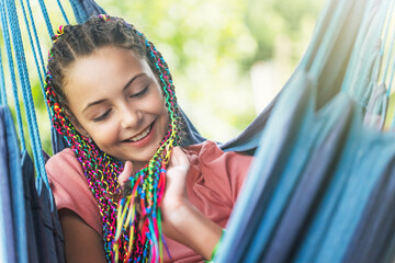 Portrait of laughing young girl with colorful braids in her hair posing on blue hammock.  Horizontally. 