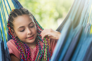 Cool young girl with colorful braids in her hair is resting on blue hammock.  Horizontally. 