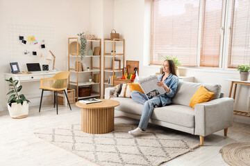 Young pretty woman reading newspaper on sofa in light living room - Powered by Adobe