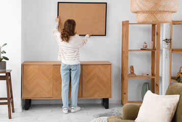 Young woman hanging picture on white wall in stylish room