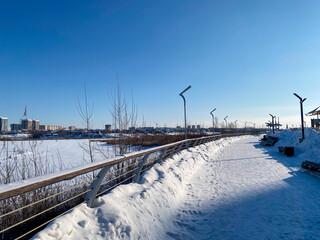 Une promenade enneigée le long d'une rivière gelée avec un pont et des roseaux, des lampadaires le long de la promenade, avec au loin la vue d'une ville