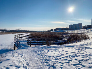 Une promenade enneigée le long d'une rivière gelée avec un pont et des roseaux, avec au loin la vue d'une ville sous le soleil