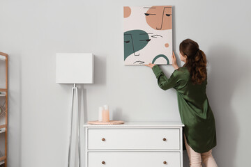 Young woman hanging picture near chest of drawers in stylish room