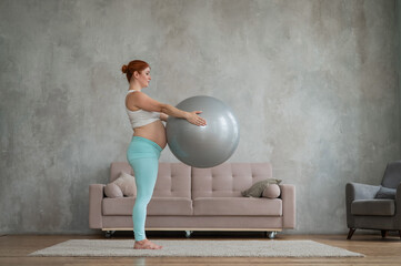 Pregnant red-haired woman doing arm exercises with fitball at home.