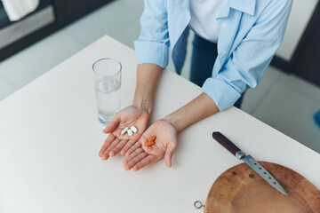 Woman holding pills and a knife next to a glass of water on a white table Concept of medication and...