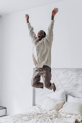 African American man jumps high on his bed, his face alight with excitement, in stylish, minimalist bedroom setting.
