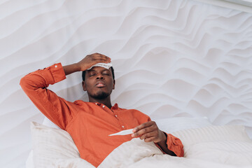African American man lies in bed appearing unwell, holding a thermometer and a cool cloth to his forehead. Young Man Feeling Unwell in Bed.