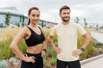 Confident couple in sportswear posing after a workout in an urban park, showcasing their fit physique and smartwatches.