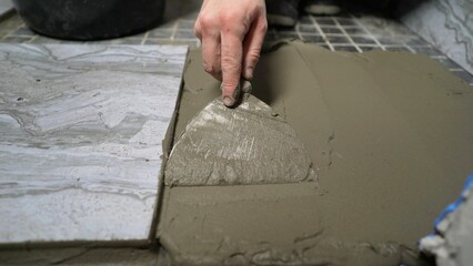  A close-up of a man's hands, in one of which he sees a spatula, with which he applies the solution to the floor. Applying adhesive - a solution for gluing tiles to the floor.