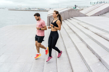 A joyful couple jogs alongside each other near the waterfront, smiling and enjoying their workout,...