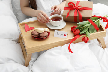 Tasty breakfast served in bed. Woman with tea, macarons, gift box, flowers and I Love You card at home, closeup