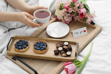 Tasty breakfast served in bed. Woman with tea, desserts, flowers and I Love You card at home, closeup