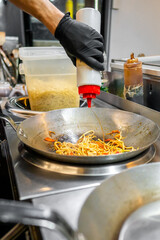 A person’s hand in a black glove cooks noodles in a wok, adding sauce from a squeeze bottle