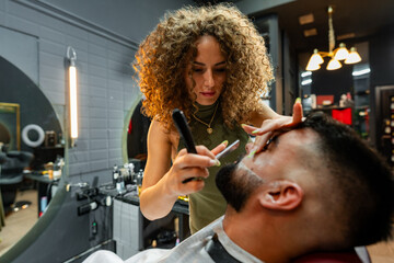 Professional barber carefully shaves and styles a man's beard using precise tools in a trendy urban...