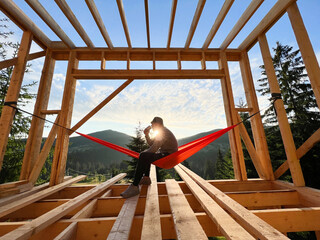 Man on construction site of wooden-framed house sits in hammock against backdrop of mountains and...