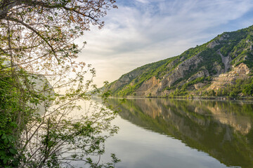 The river's calm water reflects the clear sky and the green, leafy trees that line its bank. The mountains, with their rocky terrain and steep cliffs, rise majestically in the background