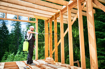 Laborer constructing wooden frame house near forest. Man treating woods, applying fire retardant using sprayer, while dressed in protective suit, helmet. Concept of modern eco-friendly construction.