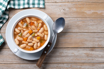 White beans soup with vegetables in white bowl on wooden table. Top view. Copy space