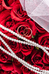 Background image for wedding design or mockup. Two gold wedding rings on a red roses bouquet background. white bridal veil. Pearl Beads. Top view.