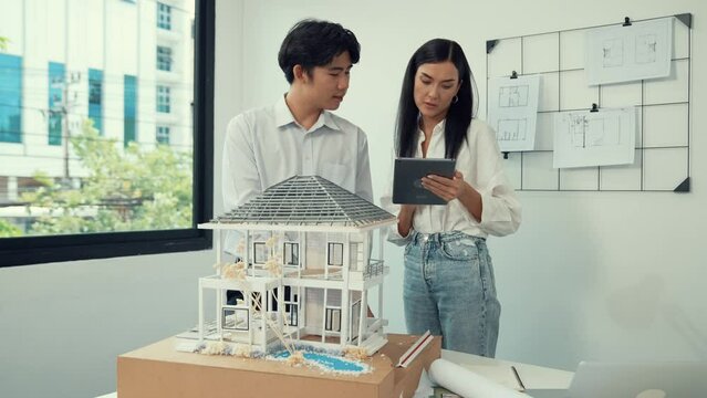 Beautiful young caucasian project manager brief asian engineer by using tablet while engineer measuring house model on meeting table with blueprint, architectural equipment scatter around. Immaculate