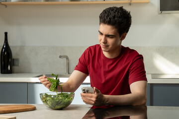 A young man in a red shirt sits at a kitchen counter with a bowl of fresh, green salad, expressing...