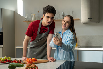 A young couple works together in the kitchen, one of them in a gray apron cuts vegetables, and the other holds a smartphone in his hands, perhaps looking at a recipe or cooking instructions, in a - Powered by Adobe
