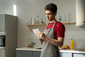 A man stands in a kitchen holding a clipboard, focused on writing down a recipe for a dish. The...