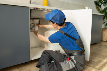 A skilled plumber in a blue uniform and cap is working diligently to repair the pipes located under...