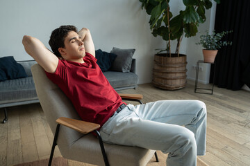 A young man wearing a casual red t-shirt and gray pants eases into a wooden chair, taking a moment...