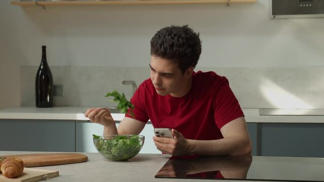 A young man in a red shirt sits at a kitchen counter with a bowl of fresh, green salad, expressing dissatisfaction with the taste. He holds a smartphone as he contemplates the unflavored dish