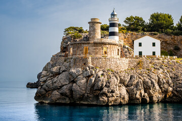 Far de sa Creu lighthouse bathed in gentle morning sunlight, graces the entrance of Port de Sóller, with old beacon ruin in foreground, capturing the beauty of Mallorca coastline and maritime heritage