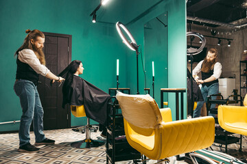 Hairdresser combs a girl .Male young hairdresser positive enthusiastic with long beard and hair in...