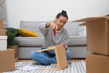 Young brunette woman installs furniture using construction tools sorting out carton boxes of things...