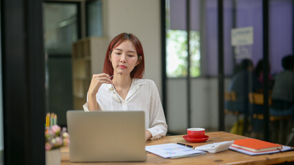 A woman is sitting at a desk with a laptop and a cup of coffee. She is wearing glasses and she is deep in thought