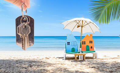 Miniature house on beach chair with hand holding house key over tropical beach background, holiday house, summer outdoor day light, property and real estate business