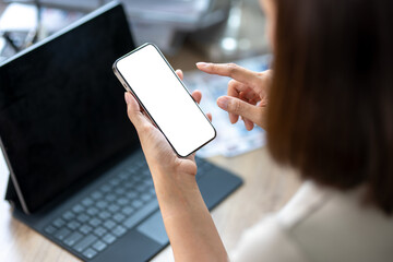 A woman is holding a cell phone and pointing at it. She is sitting at a desk with a laptop in front of her