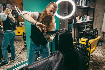 Hairdresser does a hairstyle for woman .Man hairdresser with a long beard in a vest, skillfully...