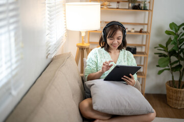 A woman is sitting on a couch with a tablet in her lap. She is wearing headphones and she is enjoying her device