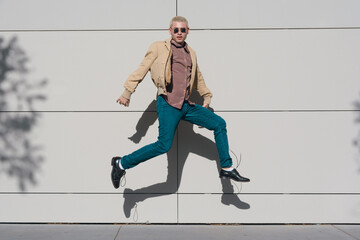 A young latin man in a tan jacket and blue jeans is jumping in the air in a sunny day