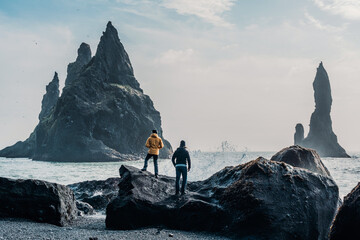 Two traveler on a huge rock on the sea. Hikers in front of the basalitic stacks of Iceland's Black...