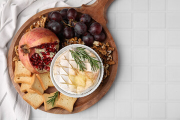 Board with tasty baked camembert, croutons, grapes, walnuts and pomegranate on white tiled table,...