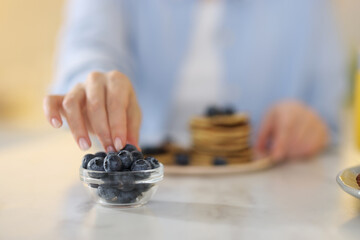 Woman taking blueberry from glass bowl at table, closeup