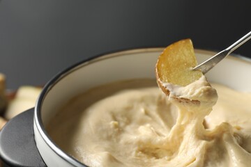 Dipping piece of potato into fondue pot with melted cheese on grey background, closeup