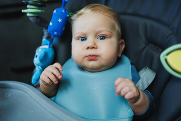 Baby wearing knitted sweater sitting in high chair and feels hungry, holding spoon and eating puree...