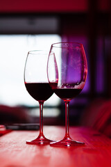 Two glass glasses with red wine inside against the light on a wooden table, indoors.