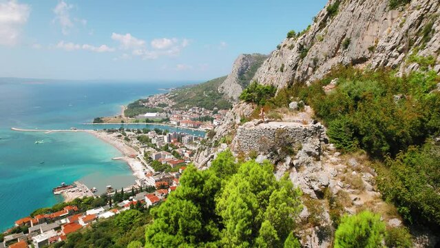 Woman looks out over a picturesque Croatian seaside town Omis from a high rocky path. Summer trekking in mountains.
