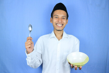 Muslim asian man wearing a cap stand and hold an empty bowl and spoon showing the dish