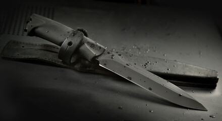 .a black travel knife and a leather sheath on a black background. There are drops of water on the...